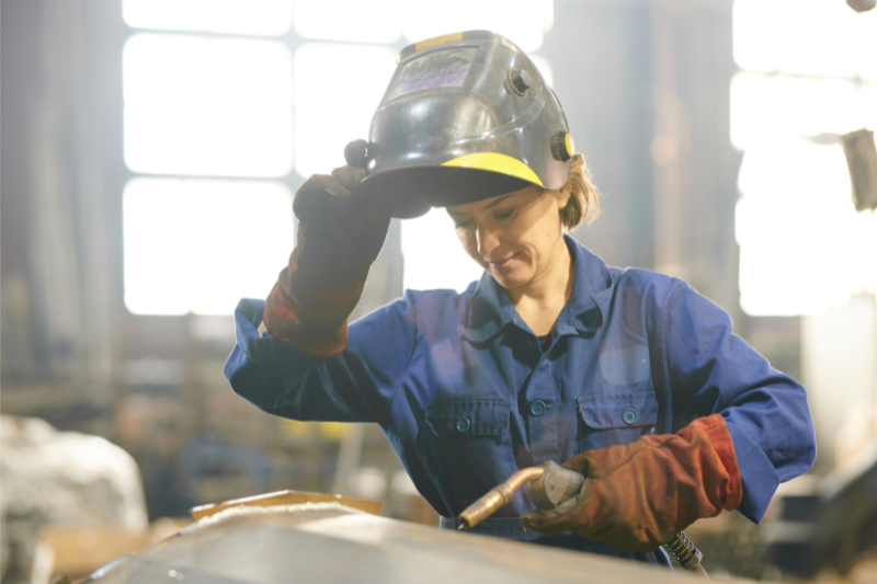 A worker in a blue protective jumpsuit raises a welding helmet with one hand while holding a welding torch in the other, standing in an industrial workshop with bright backlighting.